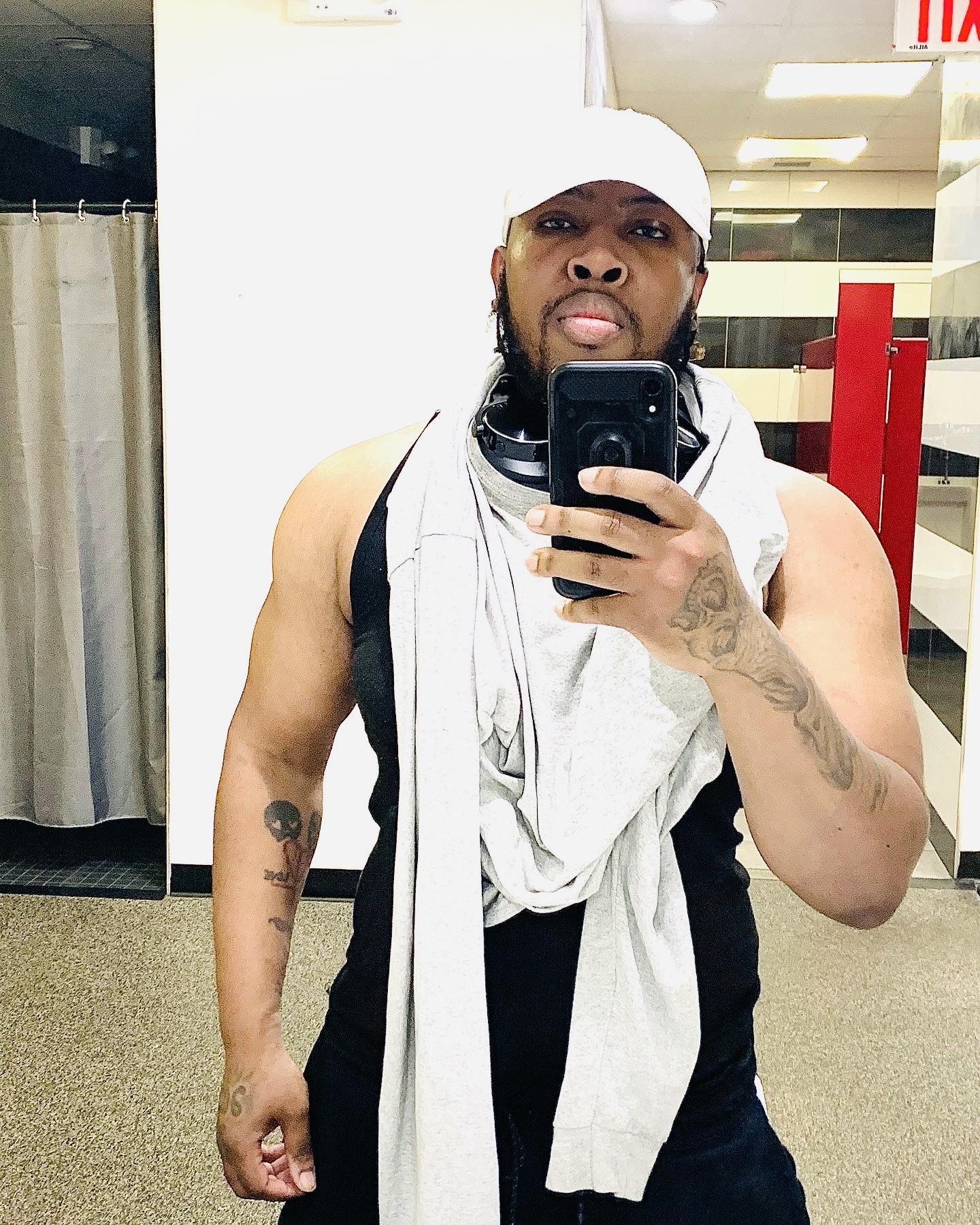 Self-portrait of the author taking a mirror selfie in a gym, wearing a white cap, black tank top, and light hoodie draped around the neck.